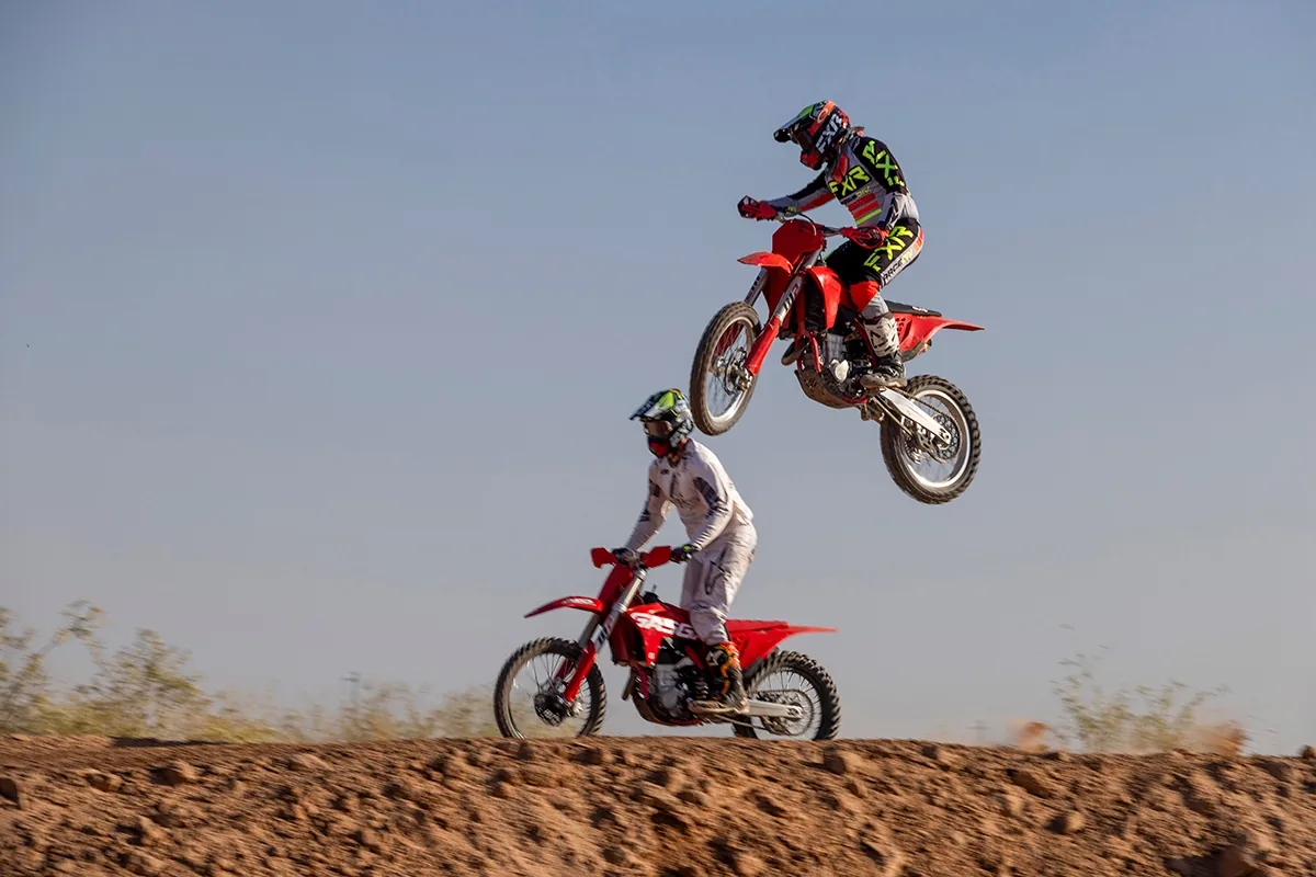 Two motocross riders catching air off a jump on a desert track